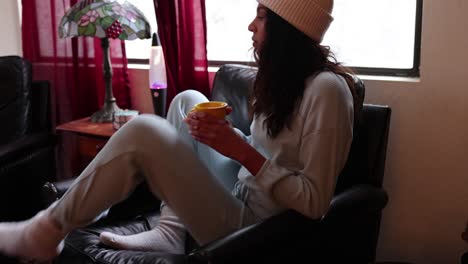 a young woman adjusts her position in a chair while she drinks hot cocoa