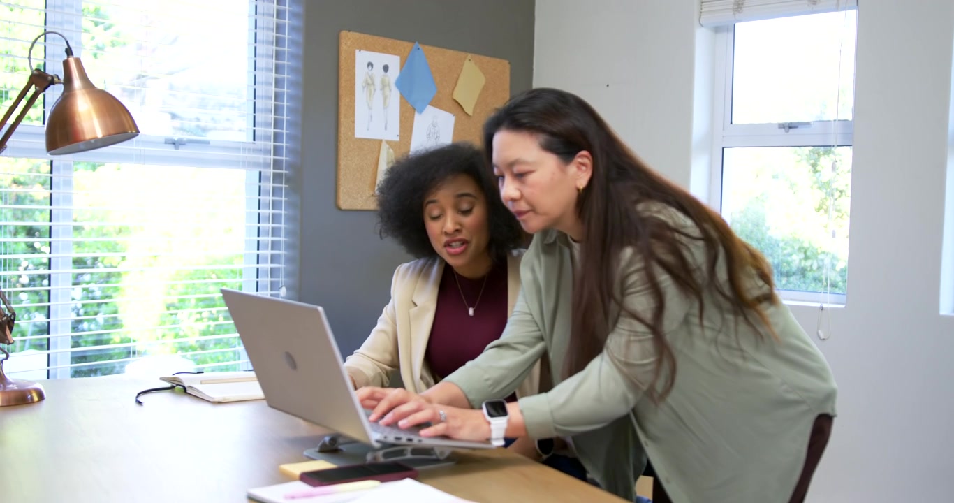 Diverse female coworkers approaching from right, leaning over laptop, typing to check sketches