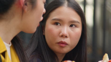 close up of two young female friends sitting on steps eating hot dogs bought at street food market stall