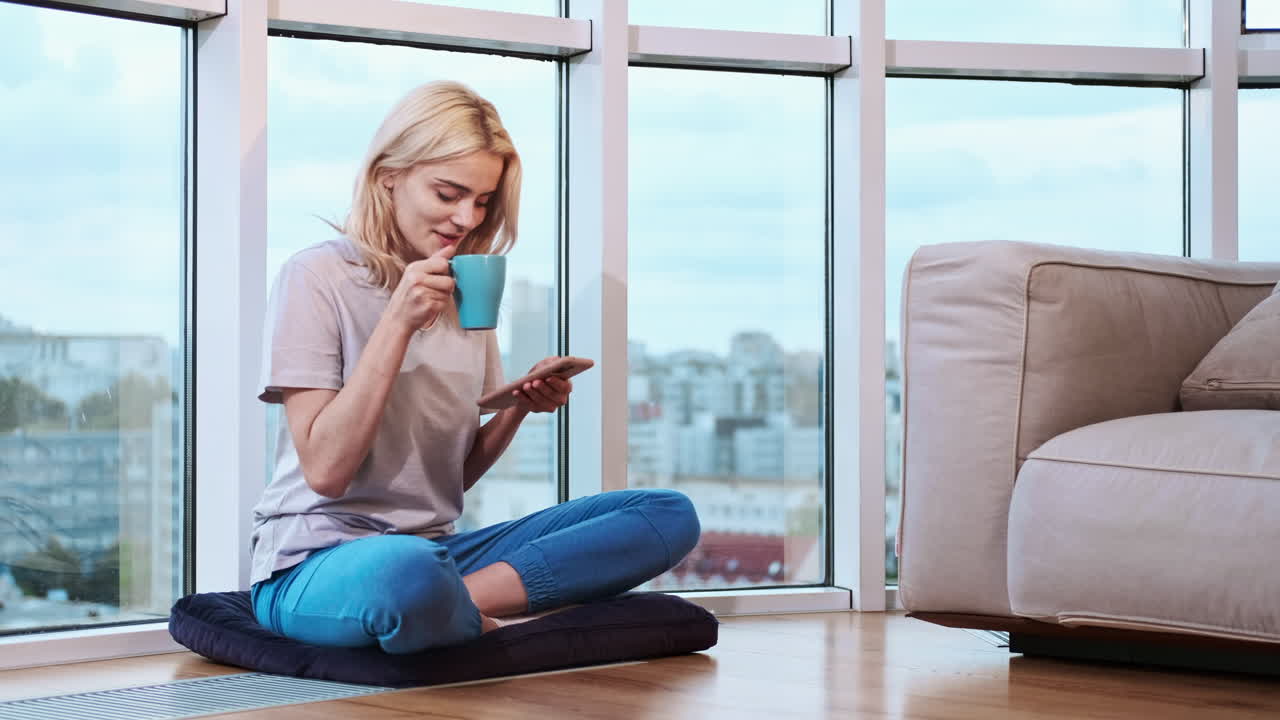 Young blonde smiling woman sitting on the floor near the panoramic window. Using her smartphone, drinking