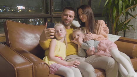 happy parents and two little girls taking selfie photo while sitting on sofa at home