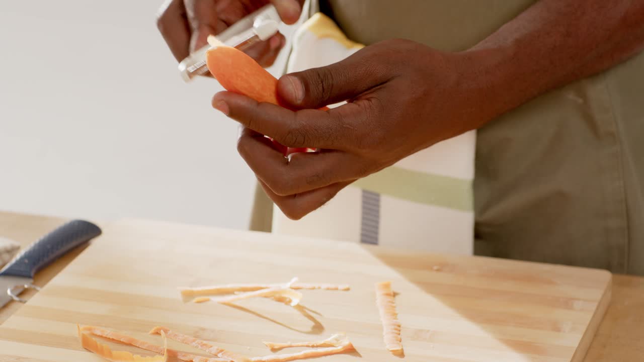 Ein erwachsener afroamerikanischer Mann in einer gestreiften Schürze bereitet eine Mahlzeit zu, indem er eine Karotte mit einem Sparschäler auf einem Brett schält.