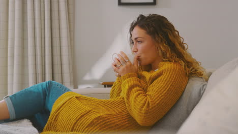 young woman in cosy warm jumper sitting on sofa at home looking out of window with hot drink