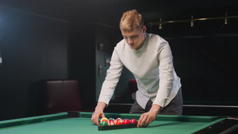 billiard trainee in white shirt and gray trousers arranges colorful billiard balls in triangle formation on vibrant green pool table under warm ambient lighting with serious facial expression