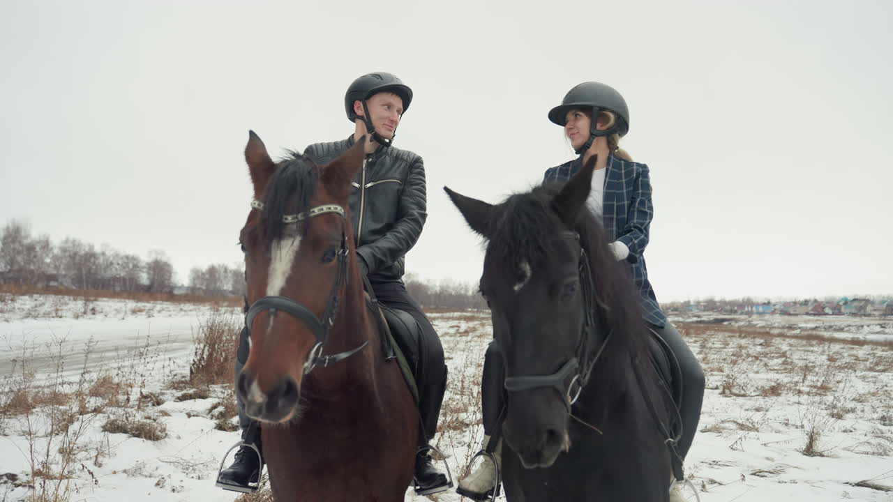 Due cavalieri caucasici su un campo innevato, scena di educazione all'equitazione in tandem con istruttore che guida lo studente, caschi e selle, trotto lento attraverso un prato invernale, conversazione calma e affiatamento.