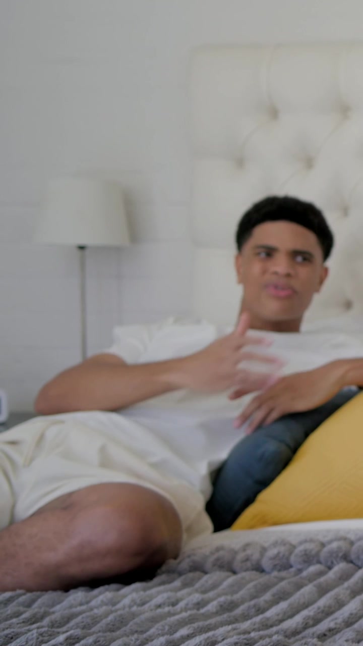 Vertical video: Hearing off screen voice, man in white tee gesturing on bed near alarm clock
