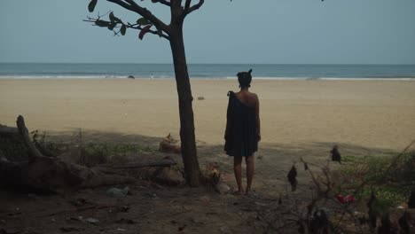 una joven solitaria está de pie junto a un árbol solitario en la entrada de una playa vacía