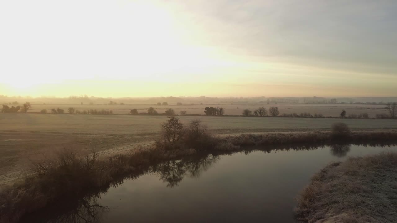 el avión no tripulado vuela bajo sobre el río serpenteante y los campos helados con un cálido amanecer a la izquierda