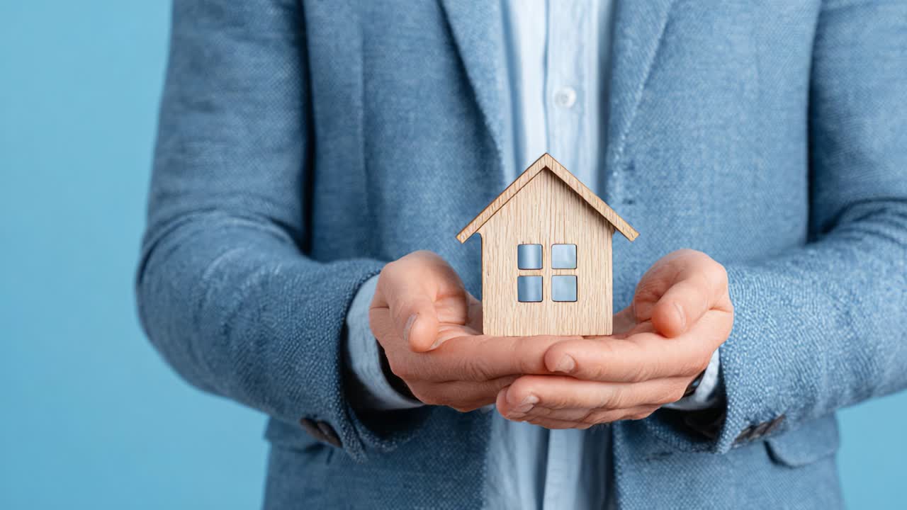 Una persona che tiene in mano un piccolo modello di casa in legno simboleggia la proprietà della casa, il comfort e il sogno di possedere una proprietà in un ambiente minimalista con uno sfondo blu tenue.