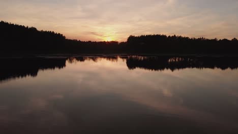 Beautiful-Sunset-By-The-Calm-Lake-Surrounded-By-Trees-In-Silhouette-In-Rogowko,-Poland---low-aerial-drone-shot