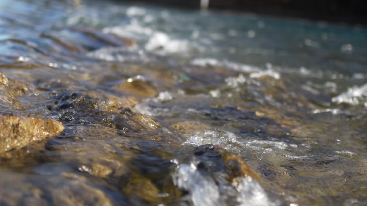 Acqua di ruscello poco profondo scorre dolcemente su rocce dorate lisce nella luce di montagna.