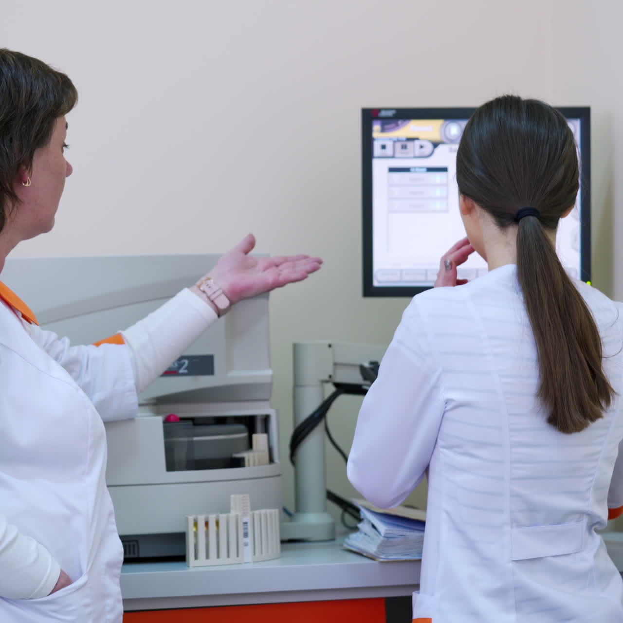 Lab workers in the medical laboratory. Backside view of two women in medical clothing looking at the screen of computer on the background of modern equipment.