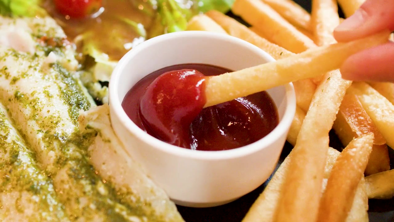 Eine goldene Pommes Frites taucht in eine Schale mit rotem Ketchup neben gegrilltem Pesto-Fisch.