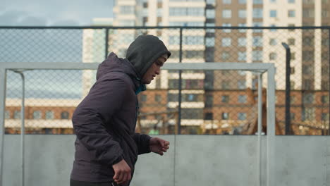 man practicing in sport arena looking visibly tired, preparing near goalpost, background features urban residential buildings and mesh fence