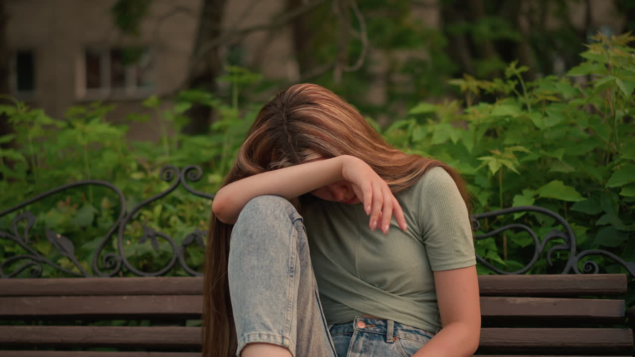 Woman in green top and jeans sits on wooden bench in park, resting her head on her arm in a reflective, somber posture, surrounded by lush greenery and trees, she conveys a peaceful solitude