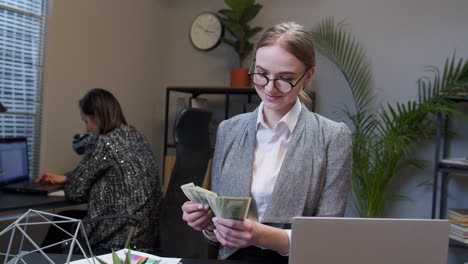 Cheerful-rich-business-woman-manager-counting-money-dollar-cash-after-working-on-laptop-at-office