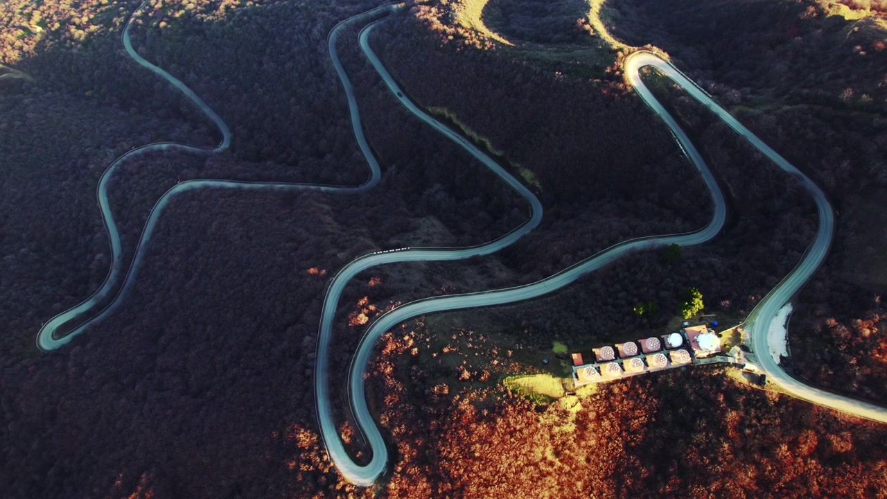Mountain road makes repeated switchbacks through leafless trees with bright curves against the dark slope while small roadside buildings line the lower junction in a high overhead view