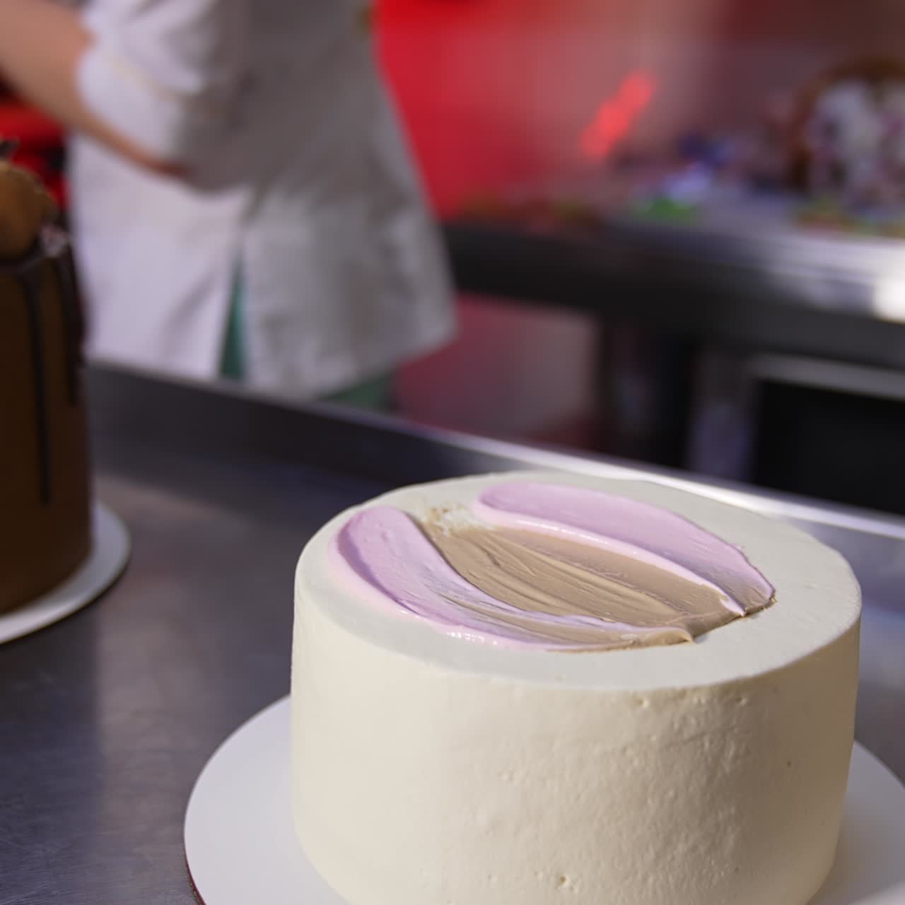 Un pastelero aplica crema blanca con una cuchara sobre un pastel blanco. Primer plano. Tarta de chocolate en la mesa al fondo
