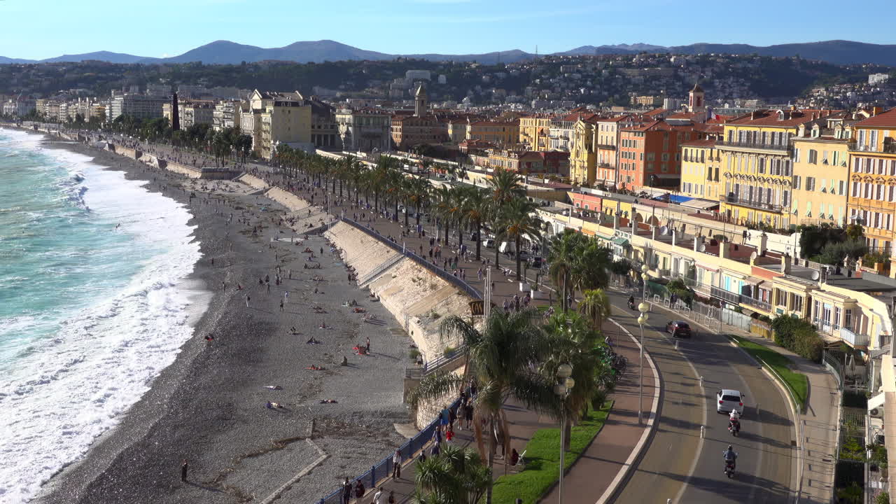 Turquoise waves crashing along the pebble beach of the Promenade des Anglais, with colorful buildings and palm trees lining the seafront