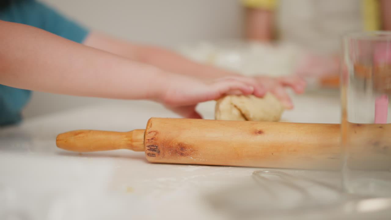 Close up of transparent glass cup placed beside wooden roller pin while little child presses dough on white kitchen table during baking session, surrounded by flour dust and baking