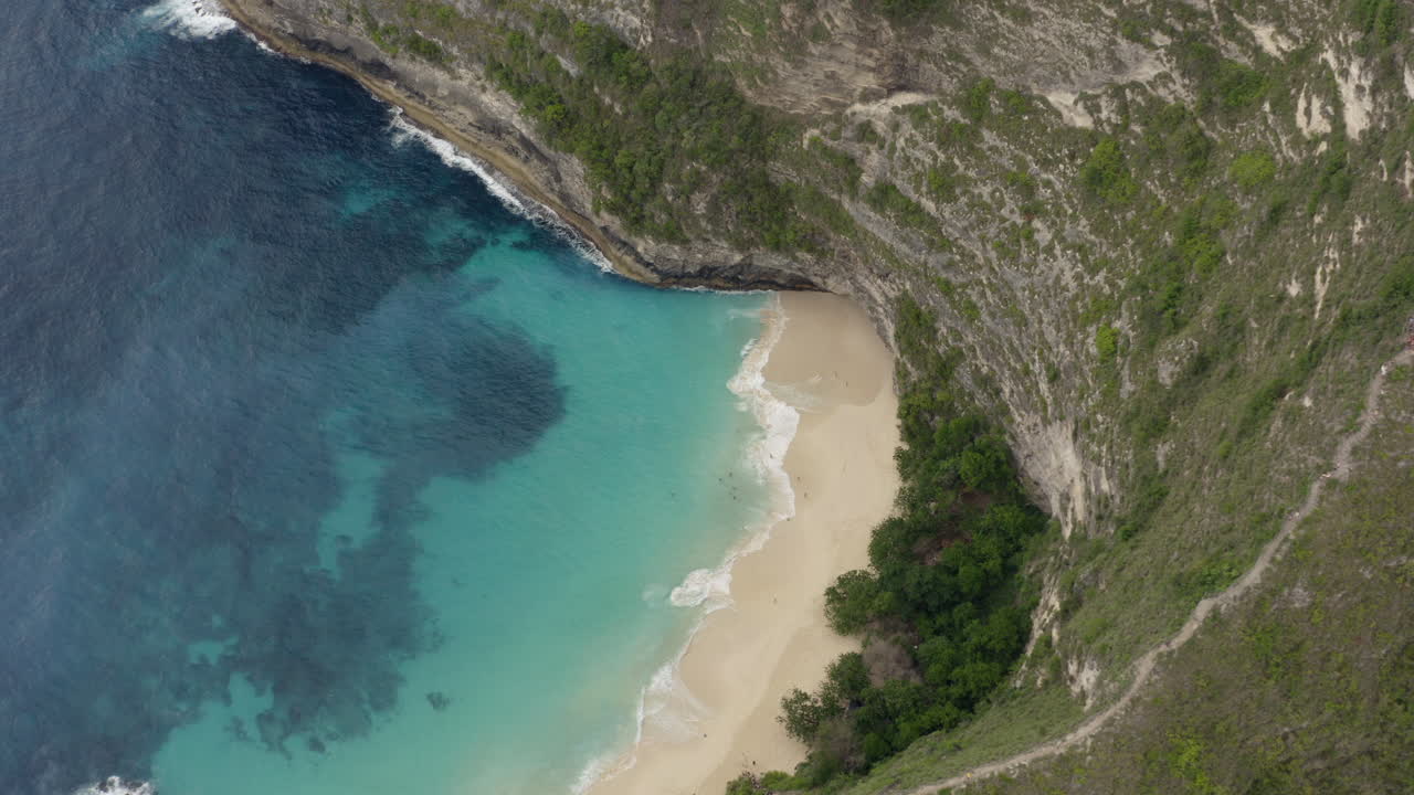 Slow pivot over Kelingking Beach surrounded by high steep cliffs with tourists walking on the sand, Nusa Penida, Bali, Indonesia