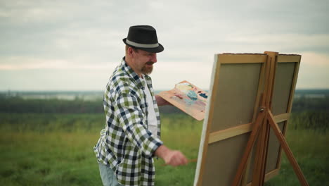 a painter, dressed in a black hat and checkered shirt, is captured mid-action, using a brush to delicately splatter paint on a large canvas in a serene grass field