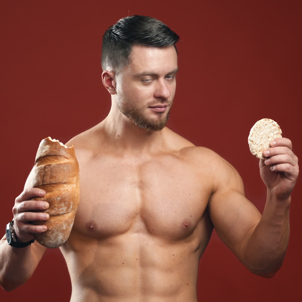 Topless sportsman with bread. Muscular athlete holding loaf of bread in one hand and a small piece of healthy product in studio. Food choices.
