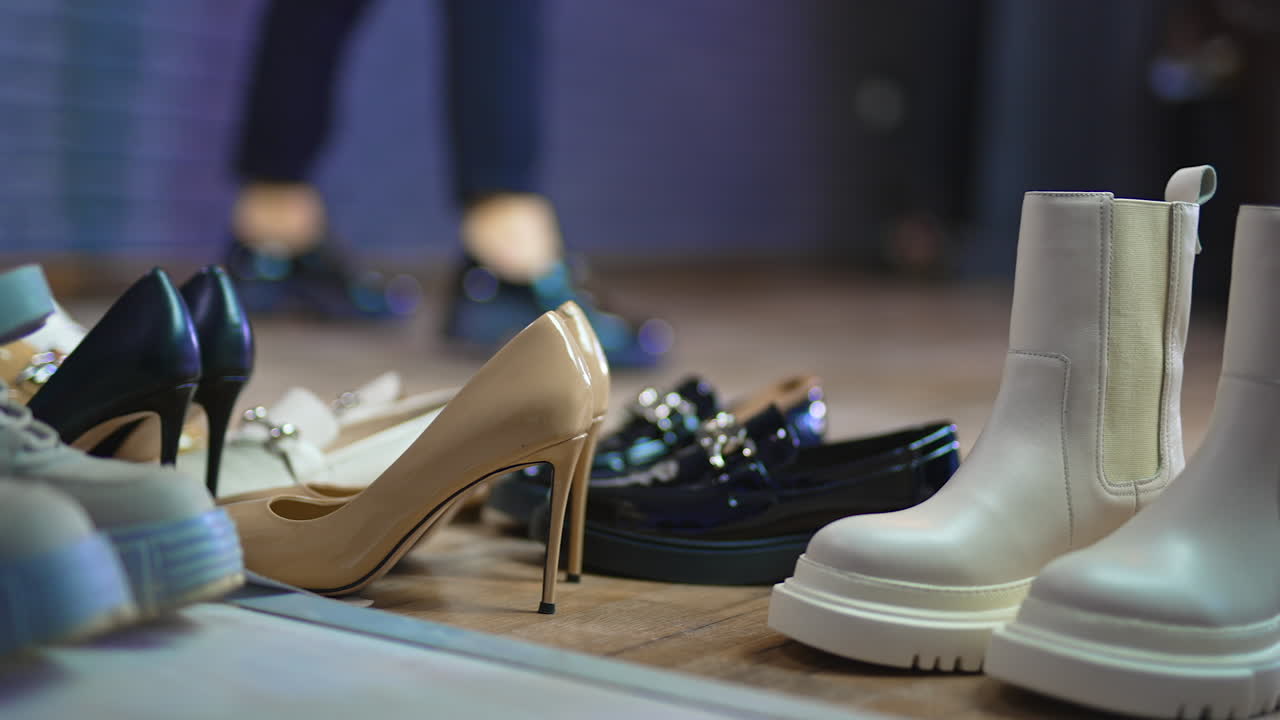 Diverse kinds of female shoes on the floor at foreground. Woman in black jeans and shoes jumping at backdrop in blur.
