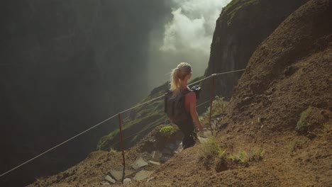 young blond woman with camera bag hiking on mountain trail along steep ridge