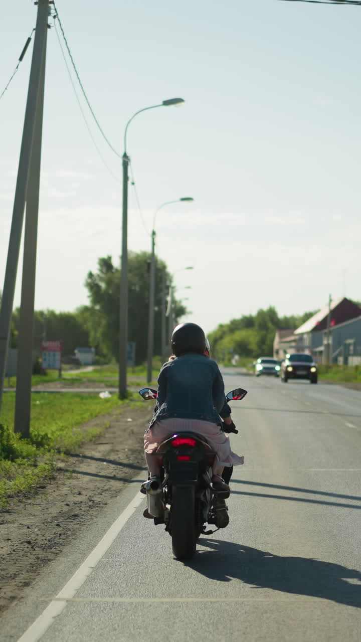 두 여인이 차분한 도로를 따라 느린 속도로 전력 자전거를 타고, 반대 방향에서 두 대의 자동차가 다가오고, 전기 기둥, 나무 및 건물이 배경에 보입니다.