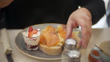 una mujer pone sal en su desayuno de bruschetta de salmón ahumado con huevos revueltos, yogur y fresas