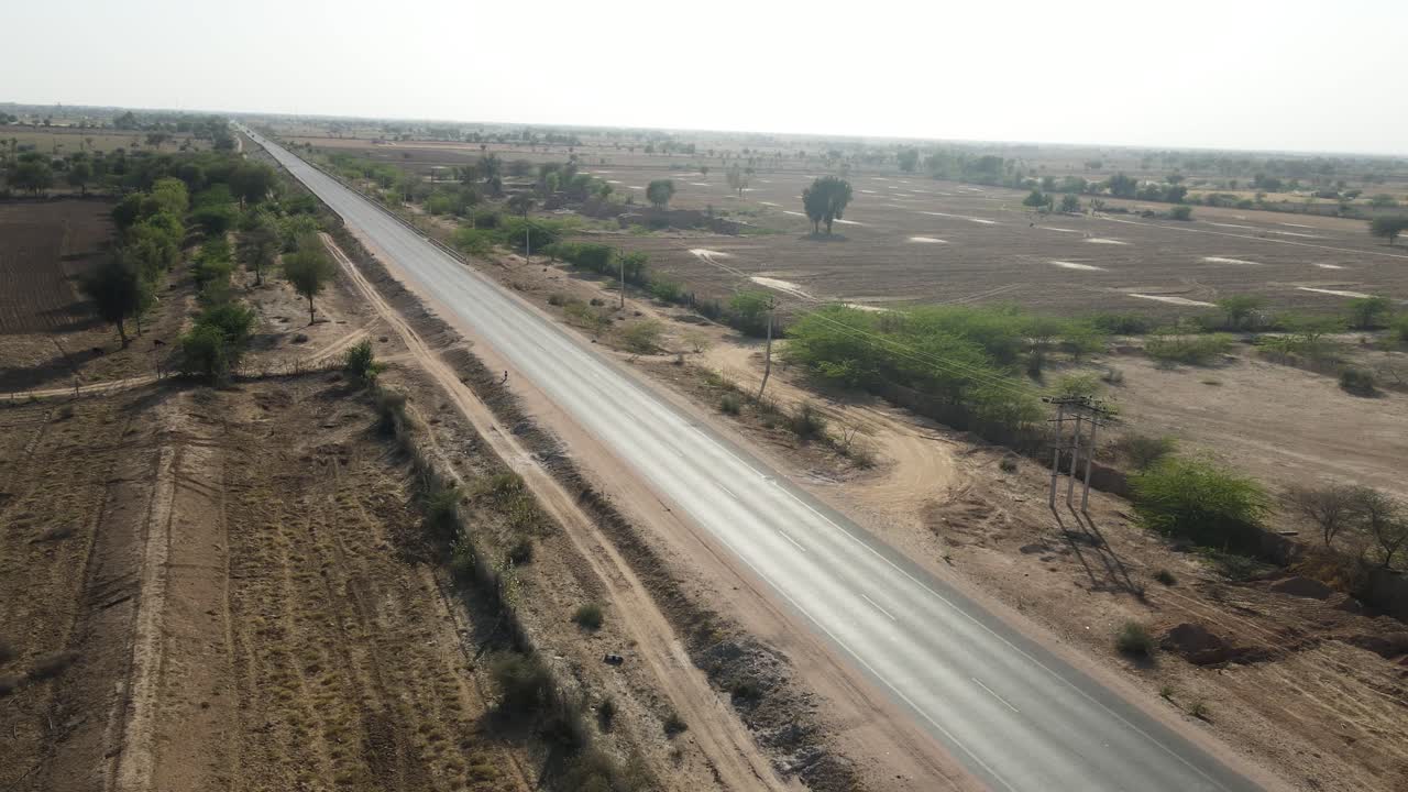 Aerial drone shot following a car on a straight road stretching endlessly through Rajasthan’s sunlit desert plains.