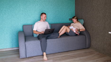 young caucasian family with a newborn baby are at home. mother is breastfeeding her child and man works at laptop.