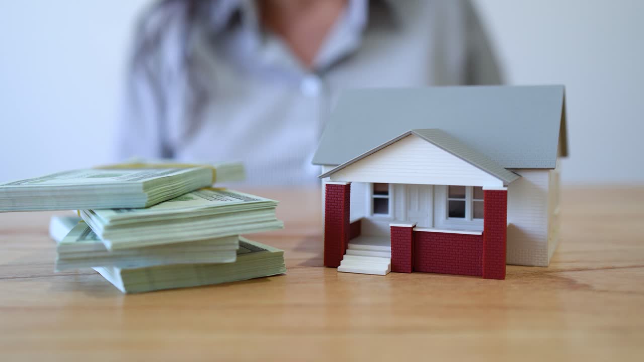 Woman Stacking Bundles Of Money With Model House - Buying Real Estate House. - closeup shot