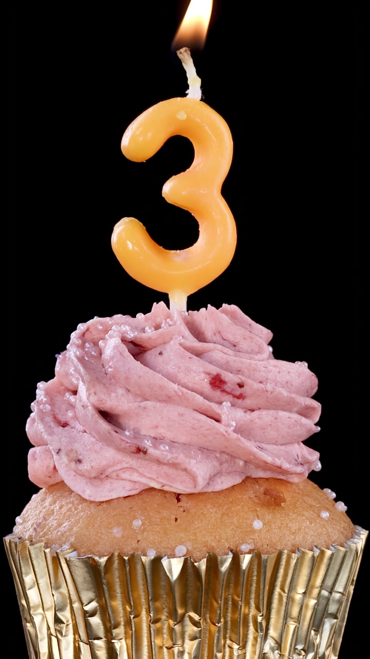 Una candela accesa con il numero tre brucia sopra un cupcake glassato alla fragola in un pirottino dorato, isolato su sfondo nero con telecamera stabile e illuminazione drammatica.