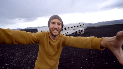 4k- young man standing by airplane wreck on black sand beach taking a selfie portrait famous place to visit in iceland and pose with the wreck