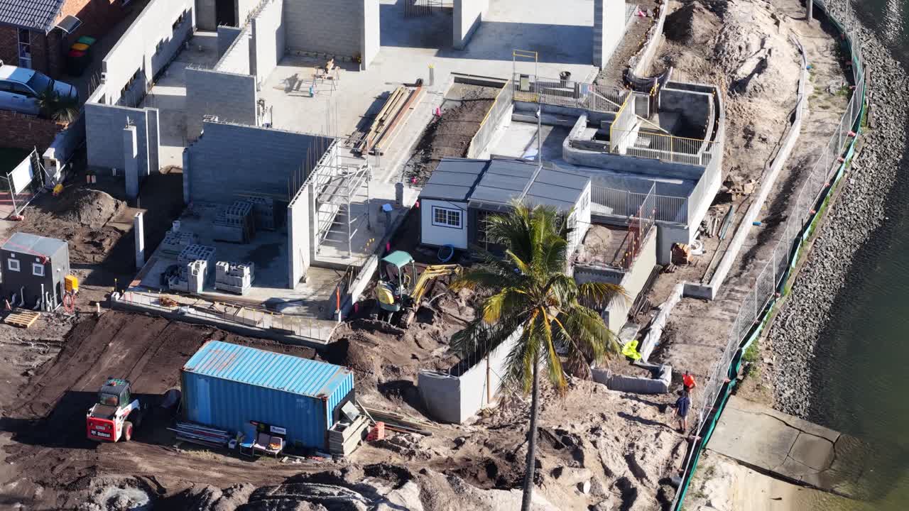 Drone footage captures construction activity at a coastal site with workers, machinery, and developing structures under bright sunlight