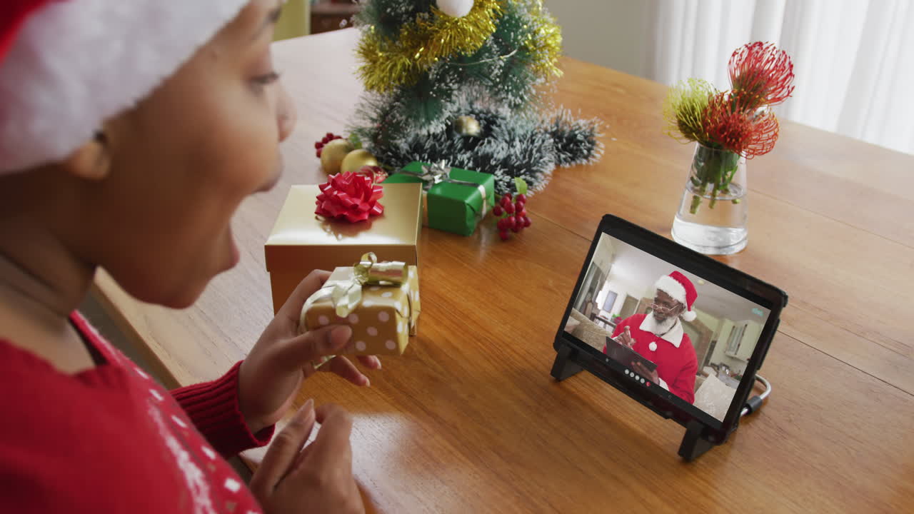 mujer afroamericana con sombrero de santa usando una tableta para una videollamada de navidad con santa en la pantalla