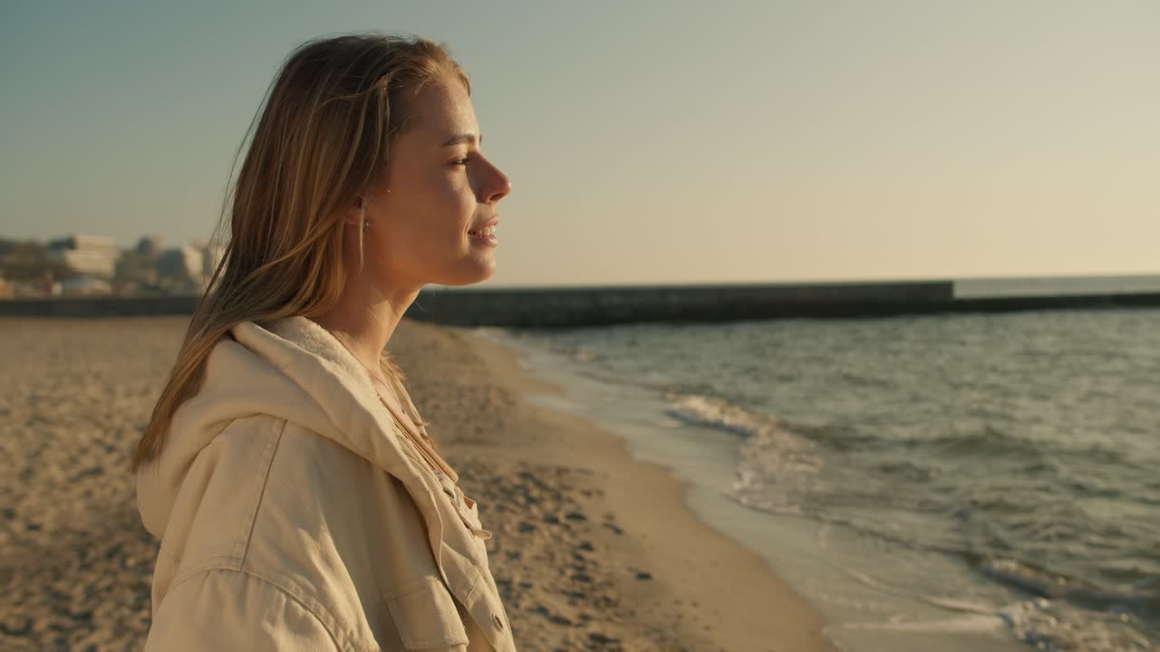 Close-up of the portrait of a young blonde girl who stands on a sunny beach in the morning looks towards the sea and smiles