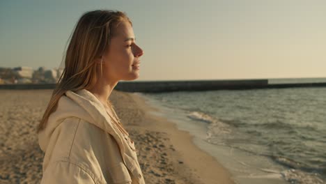 Close-up-of-the-portrait-of-a-young-blonde-girl-who-stands-on-a-sunny-beach-in-the-morning-looks-towards-the-sea-and-smiles