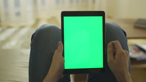 man is laying on couch at home and using tablet with green screen in portrait mode on lap
