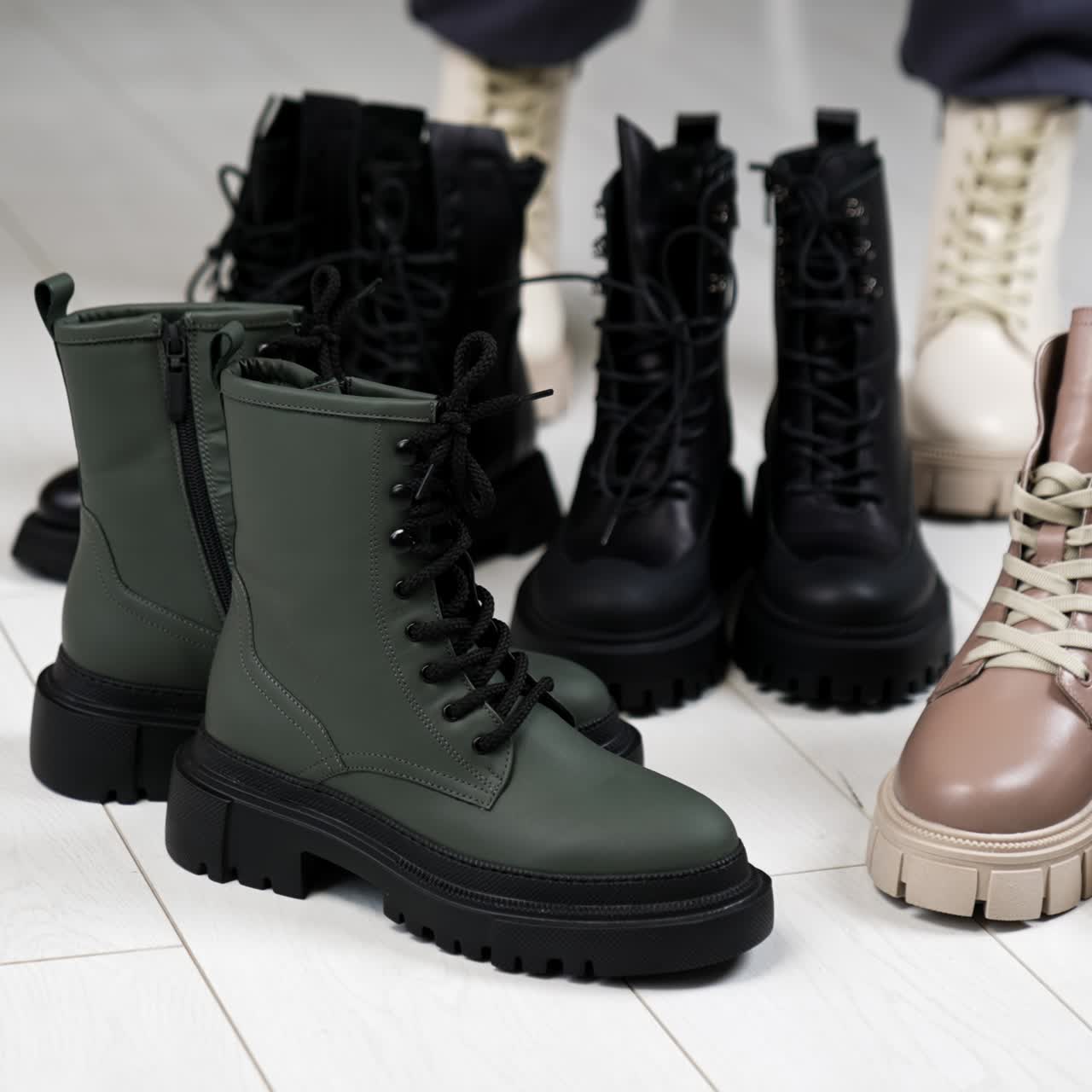 Two pairs of black and grey boots on tractor soles on the floor. Woman in white boots comes up bringing beige and black footwear