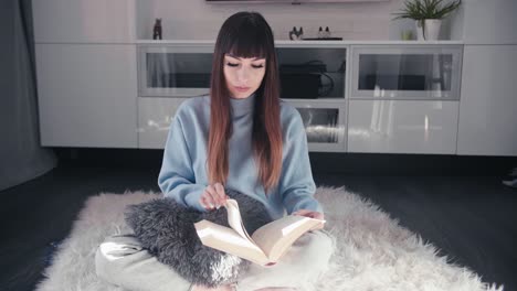 young attractive woman reading a book sit cross legged on hairy rug in her modern living room, caucasian female relaxing alone at home