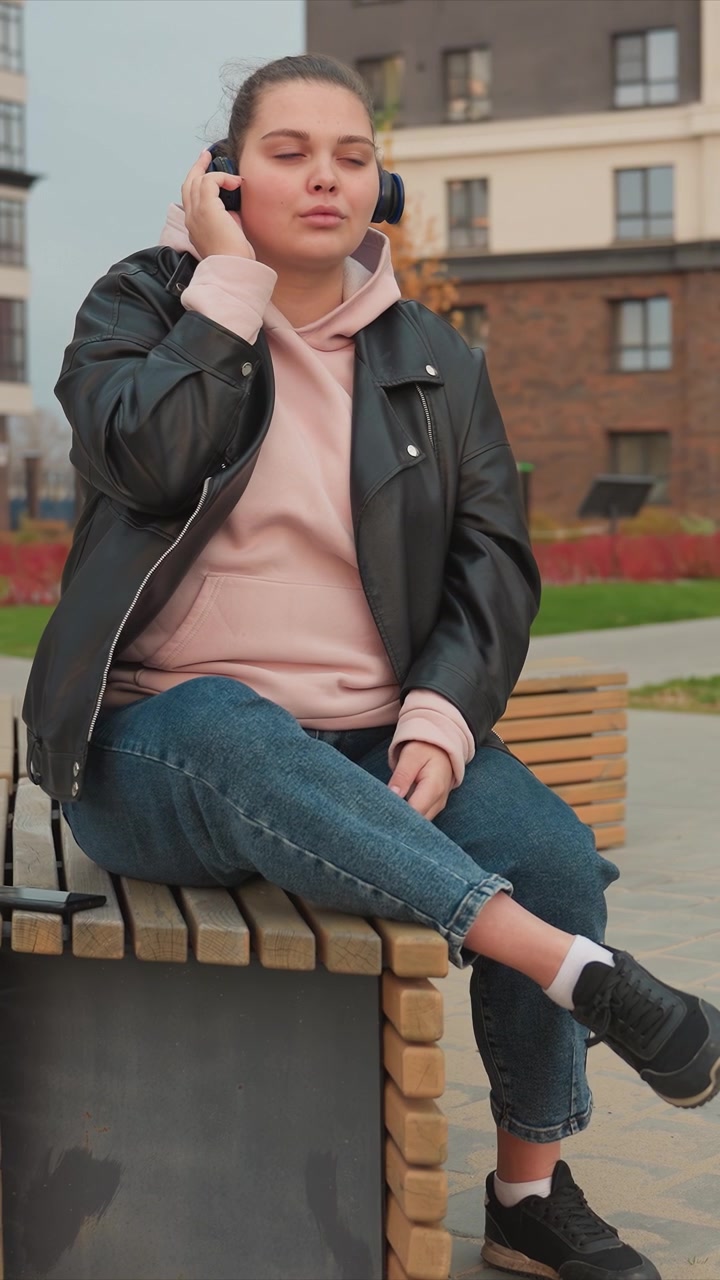Female figure adjusts earphones as she relaxes outdoors. Blue jeans rolled at cuffs paired with black sneakers. Red leaves appear near background of park like environment