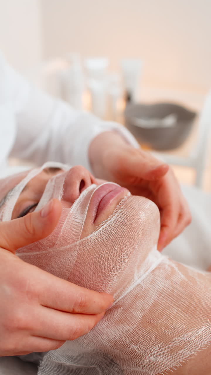 Woman treated with facial wrap mask in cosmetology clinic beautician performs massage on procedure