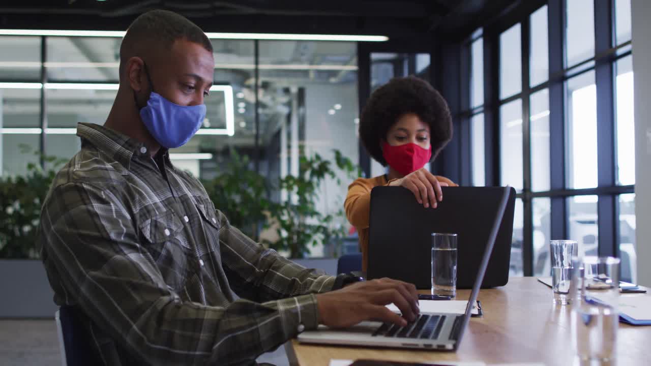 Diverse business people wearing face masks sitting using laptops going through paperwork in office