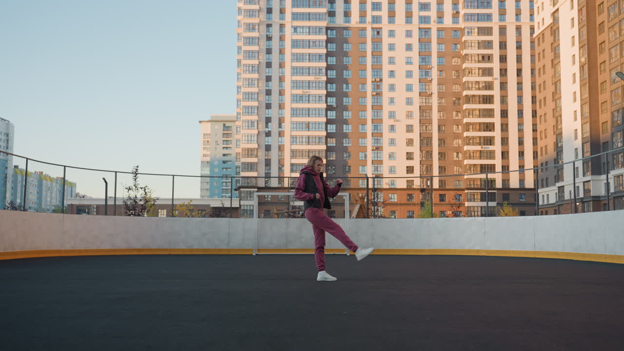 Long shot of female workout participant stretching leg inside round urban court surrounded by modern skyscrapers, wearing purple tracksuit and white sneakers under clear sky, balanced posture
