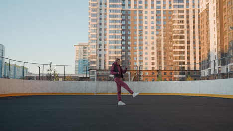 long shot of female workout participant stretching leg inside round urban court surrounded by modern skyscrapers, wearing purple tracksuit and white sneakers under clear sky, balanced posture