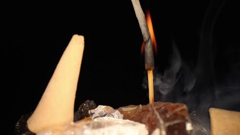 stick of incense is burning with flame all the way to end of base, wood catches fire briefly, pushing in closer to stick as flame goes out, ash bends, curls and breaks as camera pulls away