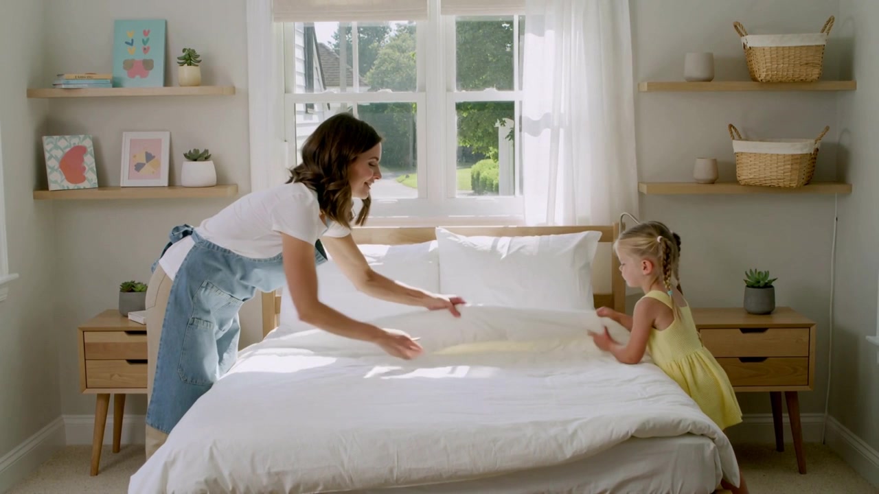 Rayos de sol iluminando a un adulto con delantal que guía a un niño con vestido amarillo mientras arreglan la cama, metiendo el edredón en el dormitorio.